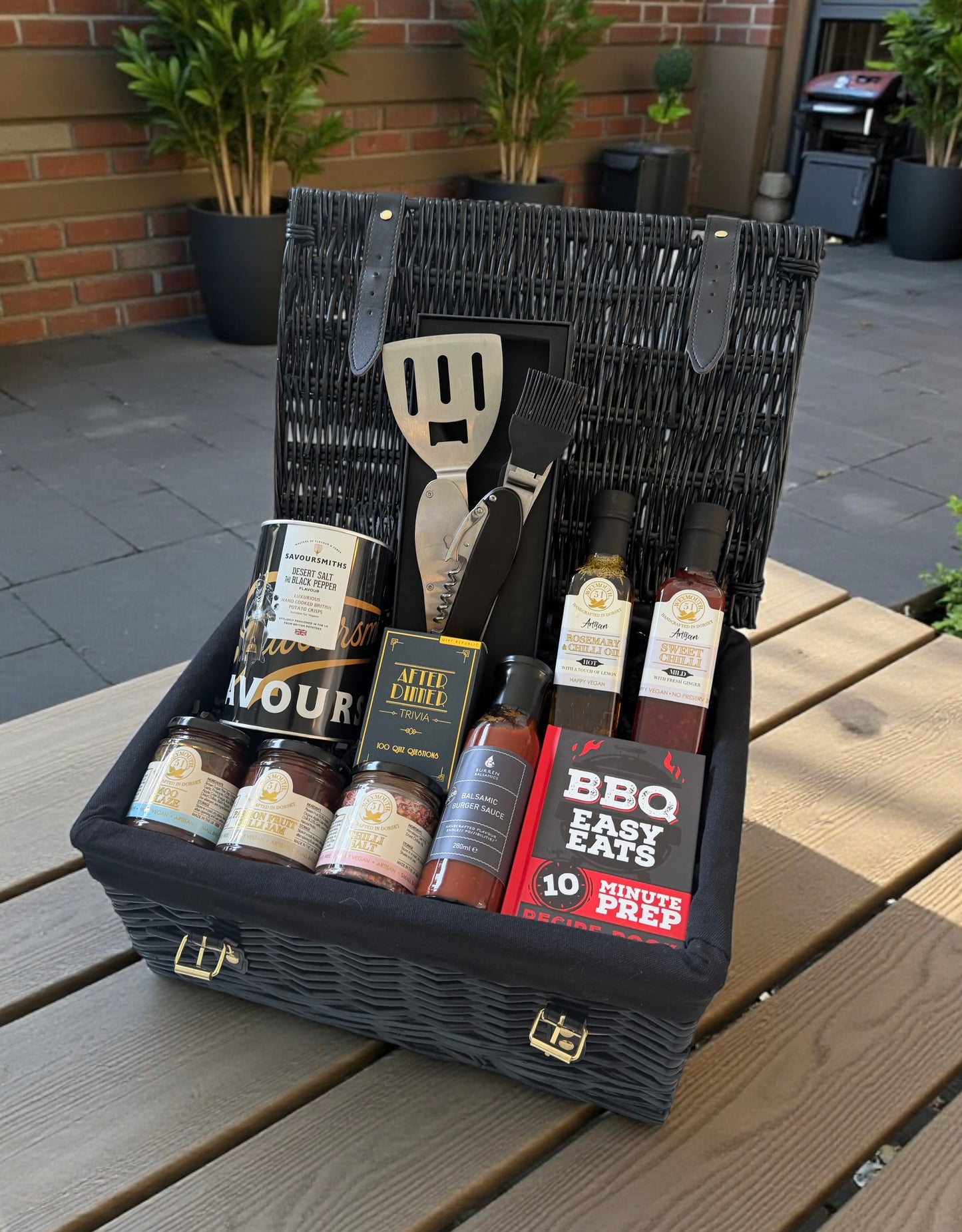 BBQ Essentials Hamper with BBQ tool, sauces, chilli oil, crisps, recipe book, seasonings, and trivia cards presented in a gift hamper.
