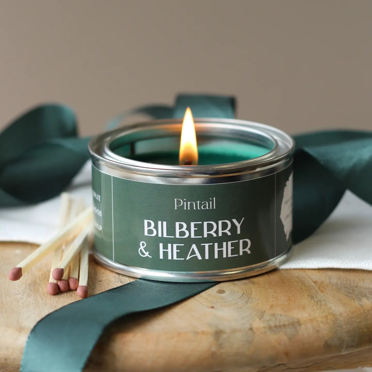 Bilberry and Heather Paint Pot Candle with a burning wick on a wooden surface, surrounded by green ribbon and matches.