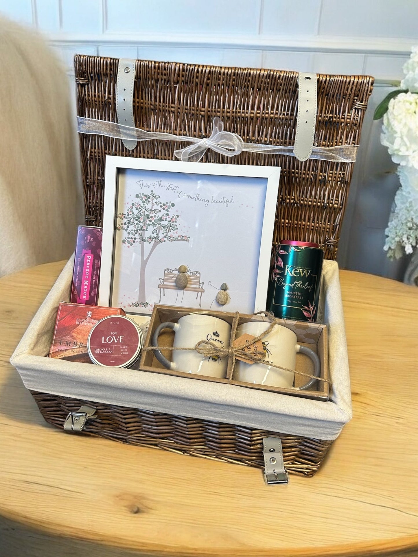 Engagement congratulations hamper with framed pebble artwork, matching mugs, chocolates, bath soak, candle, and tea caddie.