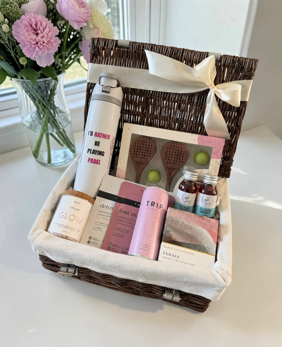 A wicker gift basket lined with cream fabric and tied with a cream ribbon, filled with assorted spa and self-care items including a white bottle, pink and white boxes, small bottles, and wooden bath tools.