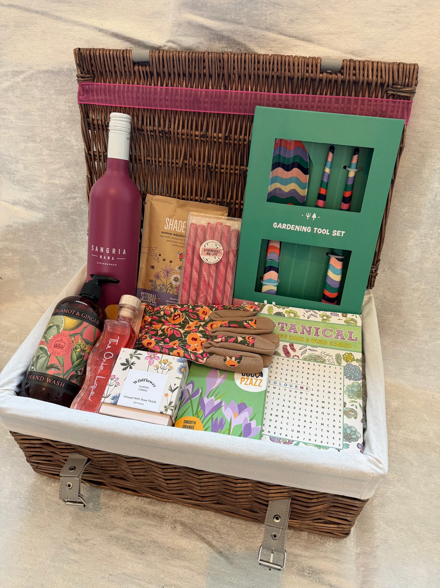 Gift basket with sangria, rhubarb liqueur, raspberry gin stirrers, orange milk chocolate, garden tools, gloves, wildflower candle, seedball pack, and hand wash, titled "Garden Sips & Scents."