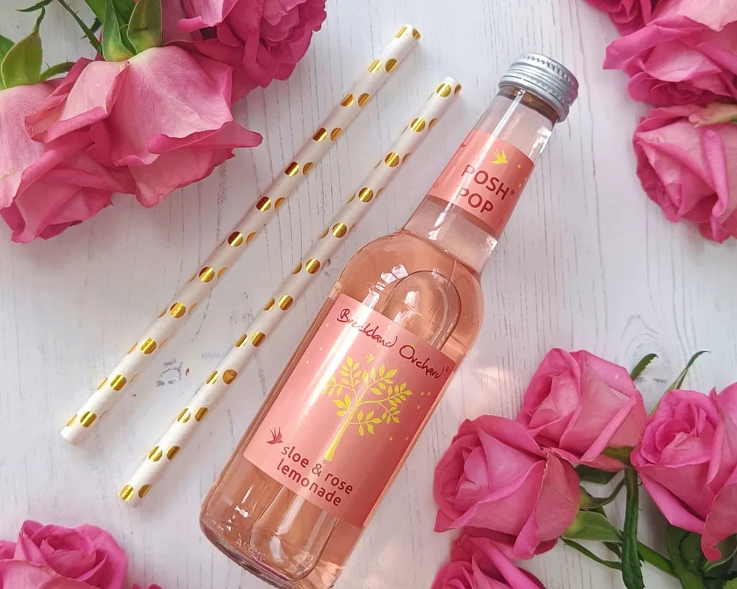 Sloe and Rose Lemonade 275ml bottle surrounded by pink roses and decorative straws, featuring a charming pink hue.