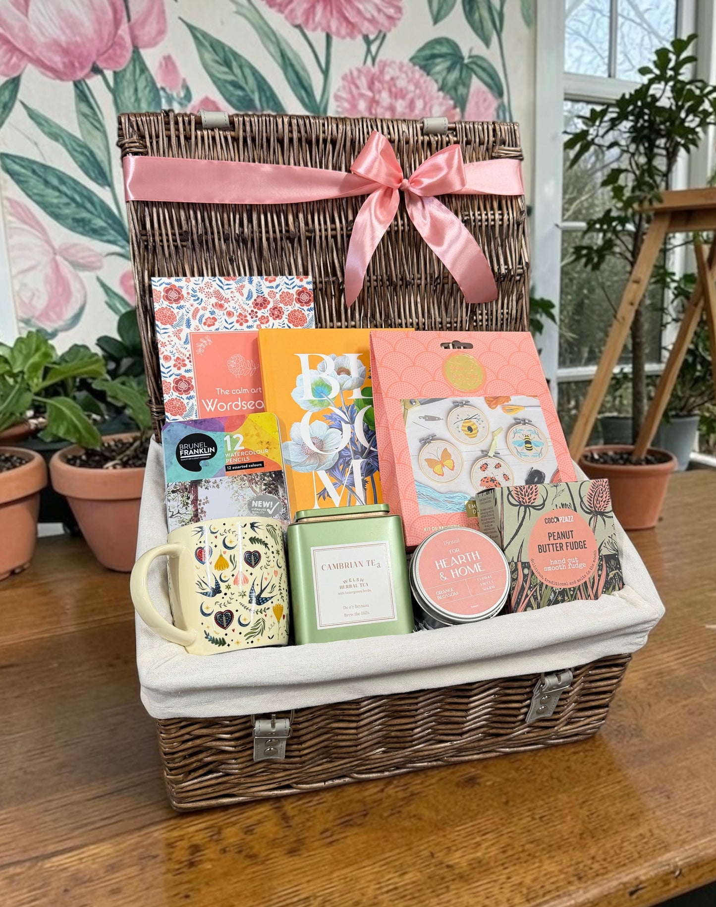 A wicker gift basket with a white fabric liner and a pink satin bow, filled with assorted gift items including a floral mug, tea, candle and sketchbook, displayed on a wooden table with potted plants in the background.