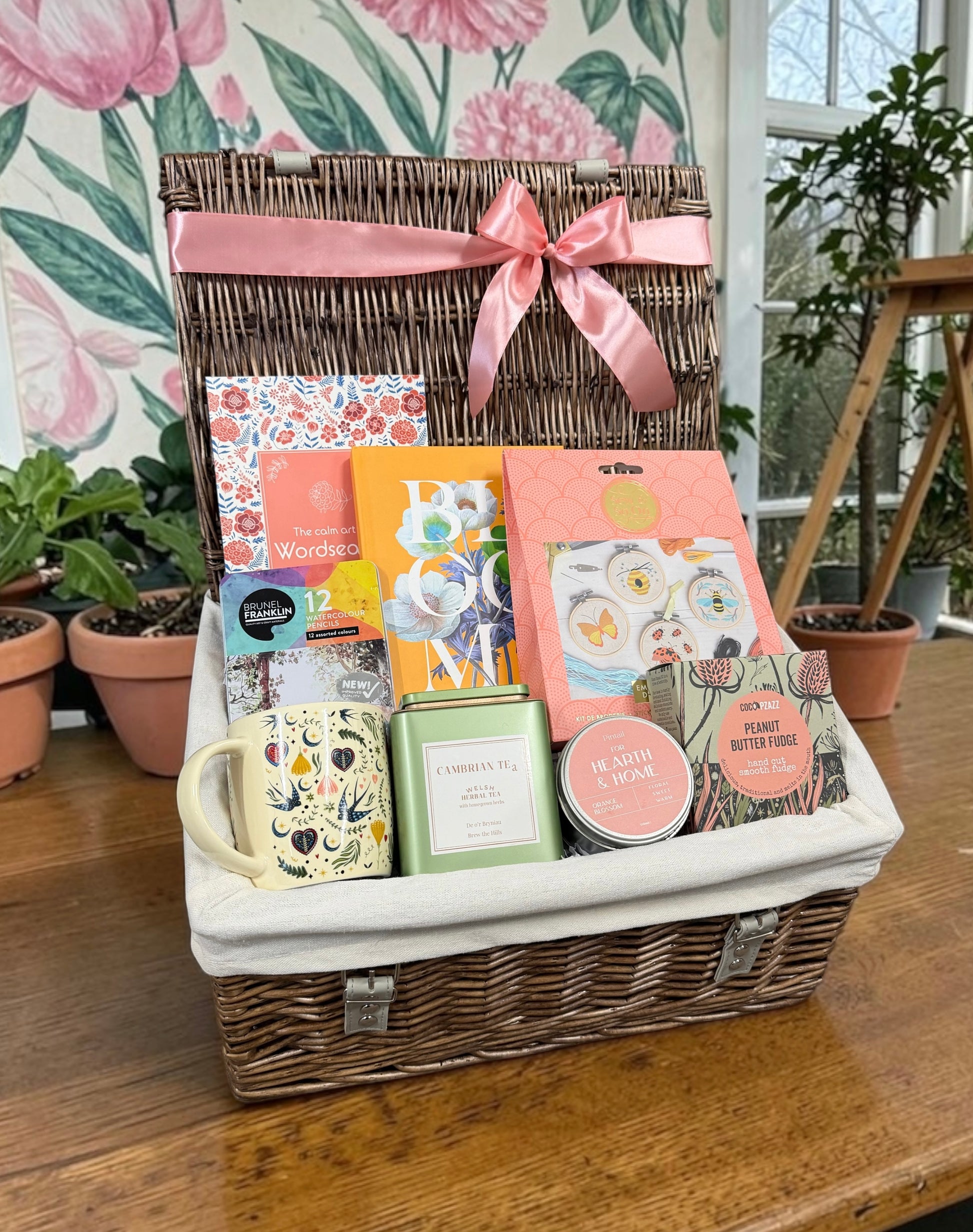 A wicker gift basket with a white fabric liner and a pink satin bow, filled with assorted gift items including a floral mug, tea, candle and sketchbook, displayed on a wooden table with potted plants in the background.