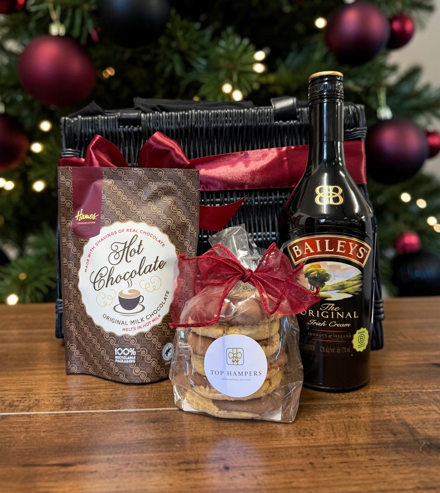 A Christmas-themed gift hamper set on a wooden table, featuring a black woven basket with a red ribbon, a bag of Hot Chocolate, a bottle of Baileys Original Irish Cream, and a bag of Tophampers cookies in front of a decorated Christmas tree.