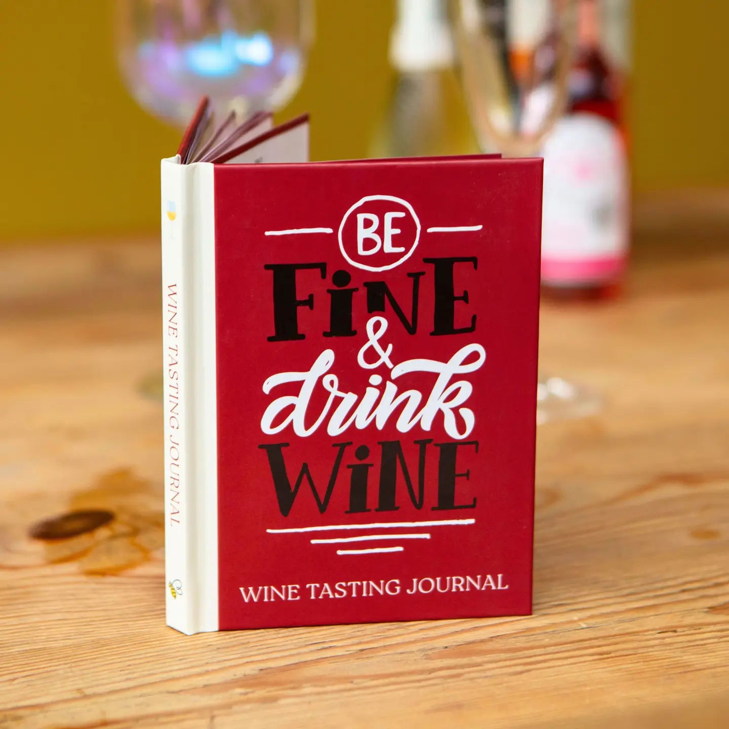 Wine tasting journal titled "Be Fine & Drink Wine" on a table with wine glasses in the background.