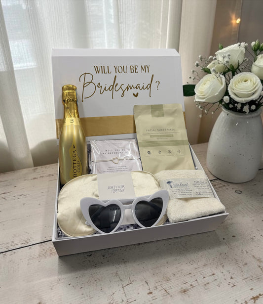 A bridal shower or bridesmaid gift box set on a rustic white wooden table, featuring a gold Bottega Veneta bottle, white heart-shaped sunglasses, a white eye mask, a pouch of white sugar scrub, a white bath towel, and a white gift bag, with a white gift box lid in the background reading 'WILL YOU BE MY Bridesmaid?'.