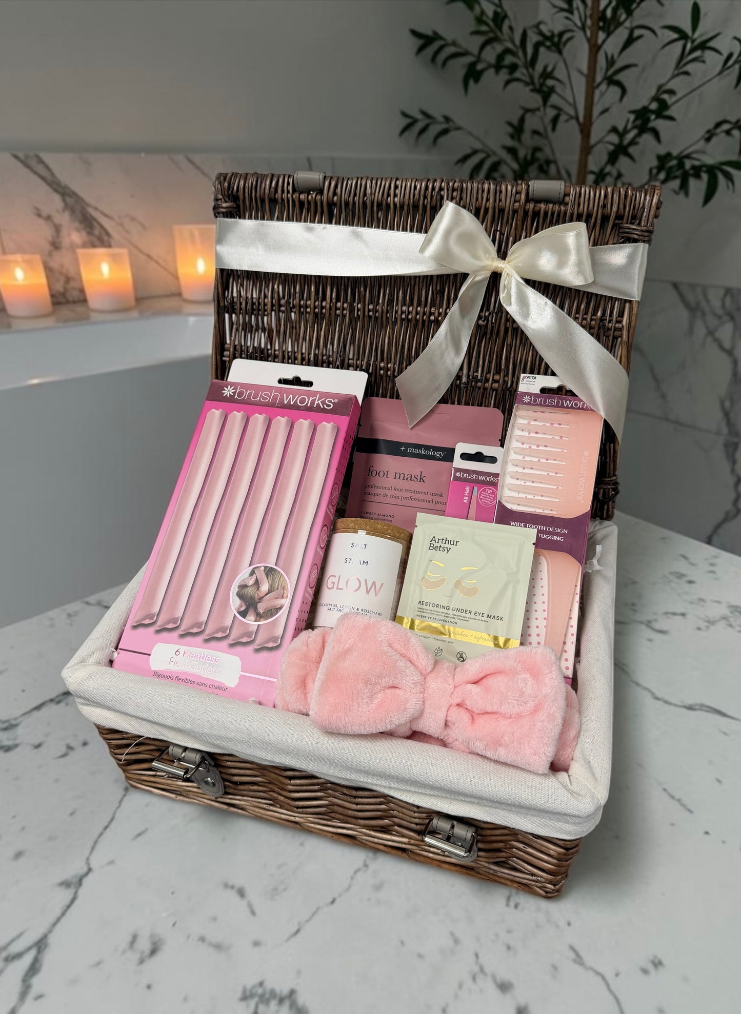 A wicker gift basket lined with white fabric and tied with a white satin ribbon, filled with pink beauty and self-care items including a pink nail set, a pink mask, a small bottle, and a pink plush bow, displayed on a marble surface with candles in the background.