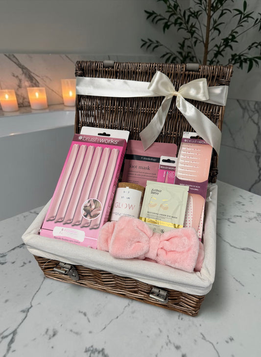 A wicker gift basket lined with white fabric and tied with a white satin ribbon, filled with pink beauty and self-care items including a pink nail set, a pink mask, a small bottle, and a pink plush bow, displayed on a marble surface with candles in the background.