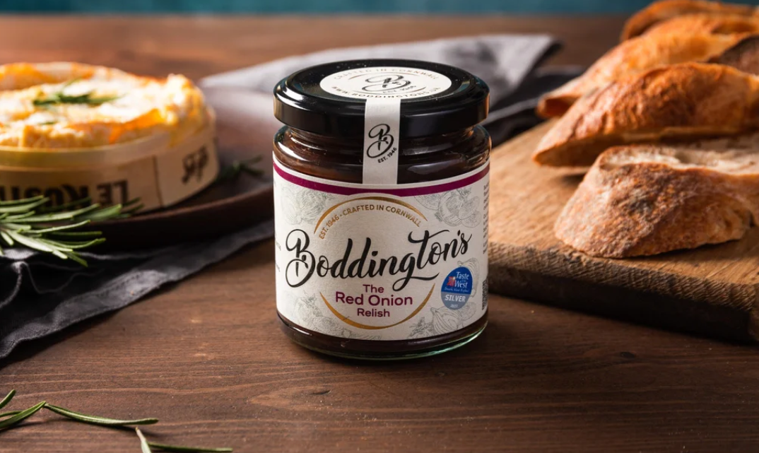 Jar of Boddington's Red Onion Relish on a wooden table with bread and herbs.