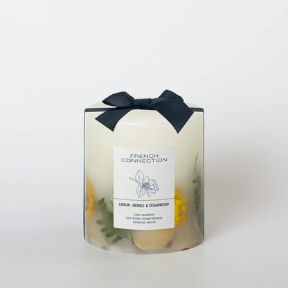 A white candle with clear elements at the bottom, infused with botanicals, wearing a black bow on top, with a label that reads 'FRENCH CONNECTION' and the scent 'LEMON, NEROLI & CEDARWOOD'.