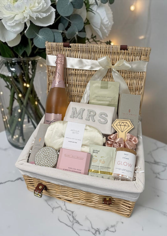A wicker gift basket filled with bridal and self-care items, including a pink champagne bottle, self care items, a MRS make up bag, skincare packets, and a satin eye mask 