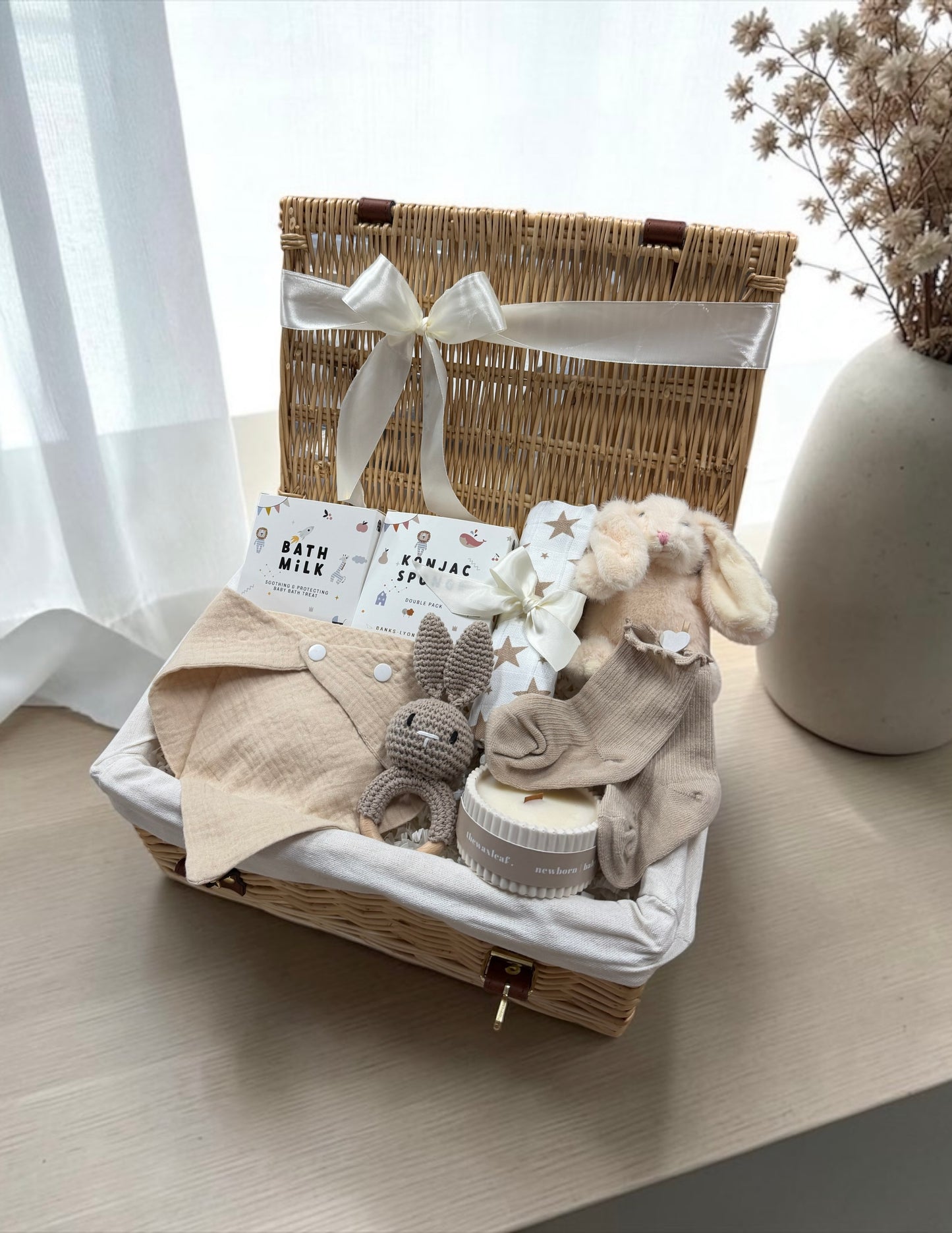 A baby gift basket set in a natural wicker picnic basket with a white satin ribbon, containing a beige quilted baby romper, a knitted bunny toy, a plush bunny, a small jar, and bath and body packets, displayed on a wooden table next to a vase of dried flowers.