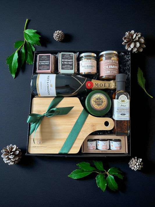 Champagne Cheese Board featuring Moët Champagne, vintage cheddar, pigs in blankets nuts, cumin olives, rosemary & chilli oil, miniature preserves, and paddle serving board.