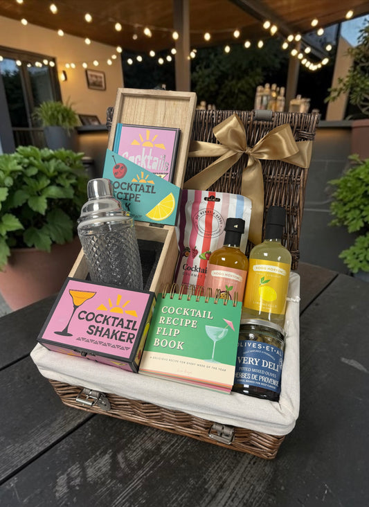 A wicker gift basket filled with cocktail-themed items, including a shaker bottle, a cocktail recipe flip book, a cocktail shaker box, and a decorative ribbon, displayed on a dark outdoor table at night with warm string lights in the background.