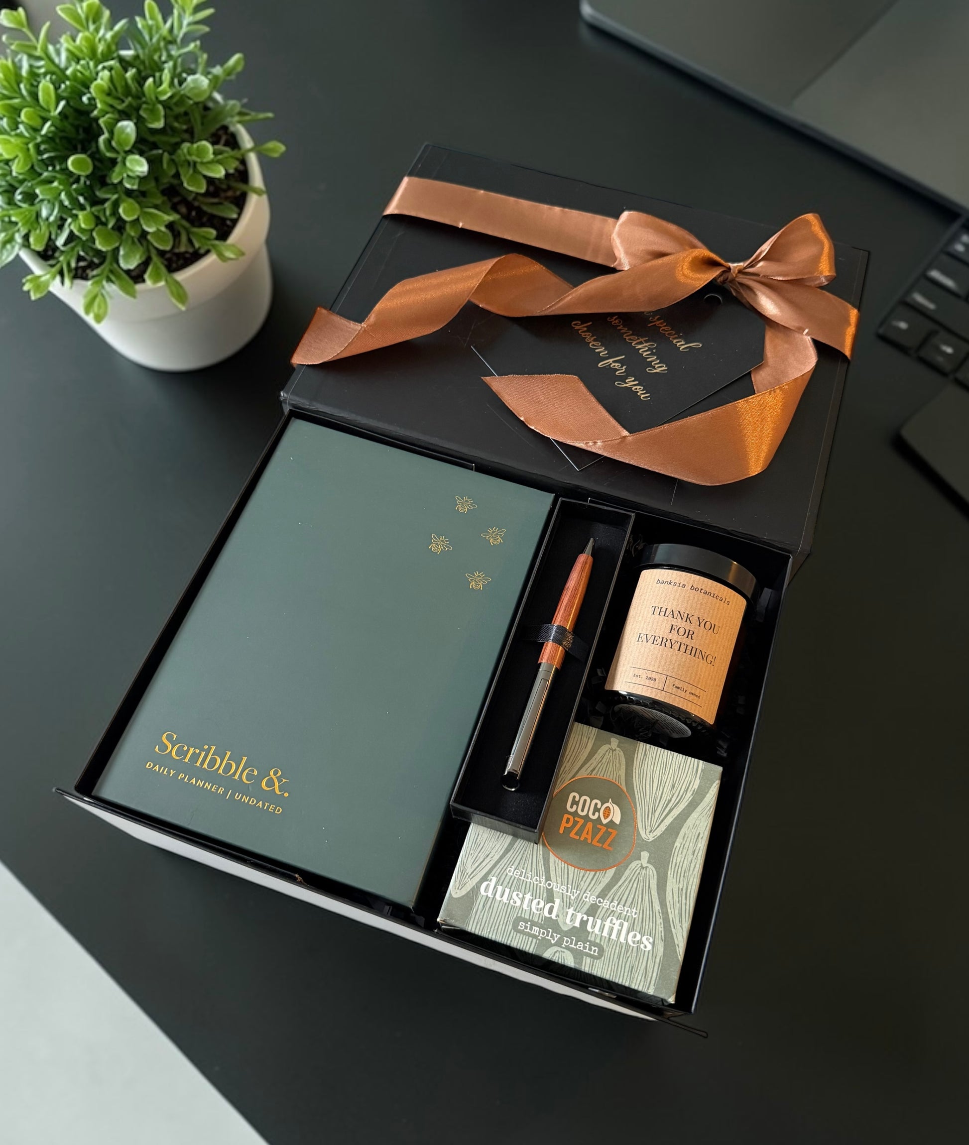 Thank you Gift set with a candle, pen, journal, chocolates  and other items on a desk with a plant and laptop.