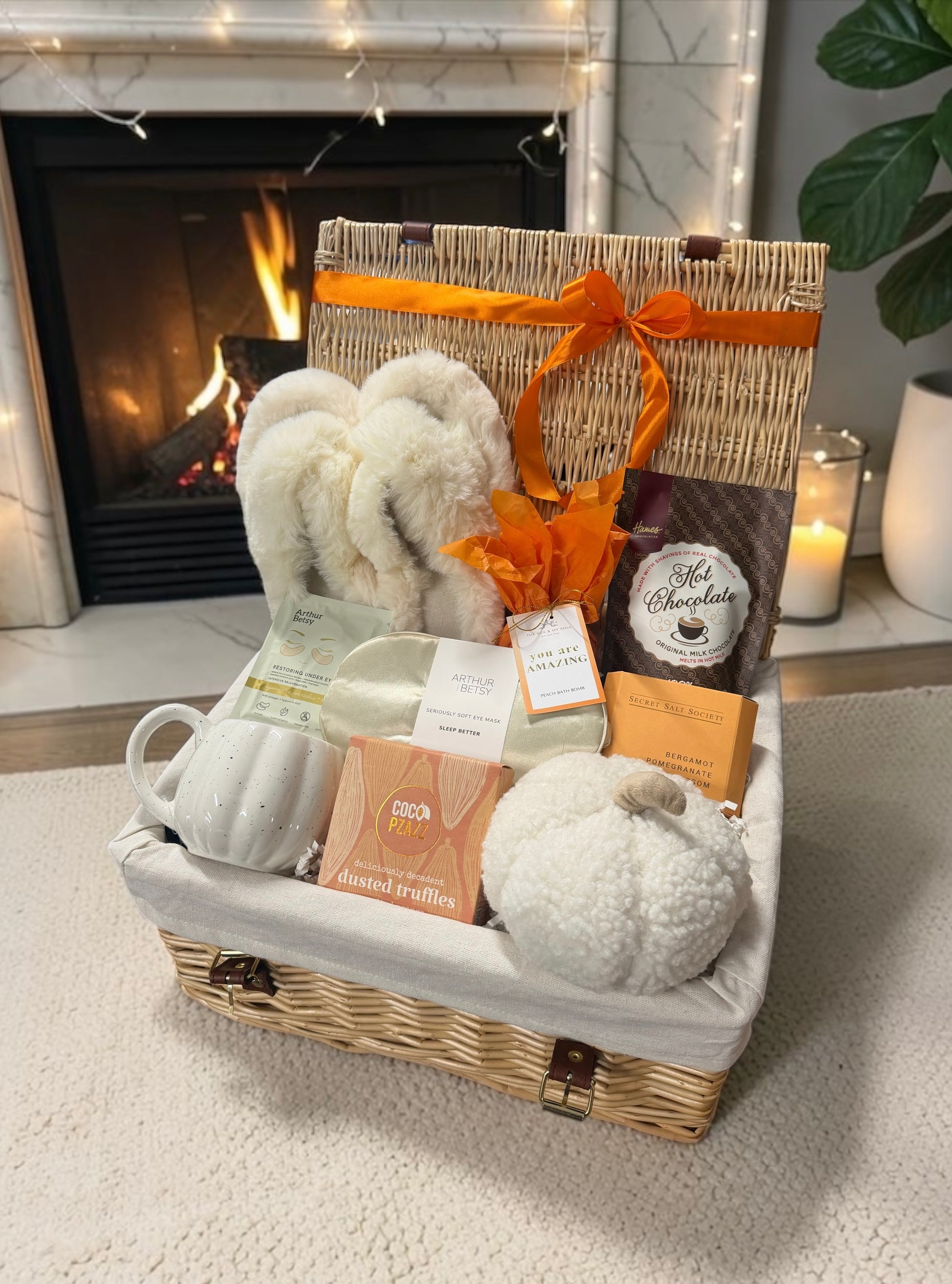 A wicker gift basket filled with autumn and comfort items, including a white ceramic mug, fluffy white slipper, a pumpkin-shaped soft plush doorstop, hot  chocolate, and a bath bomb set in front of a lit fireplace. 