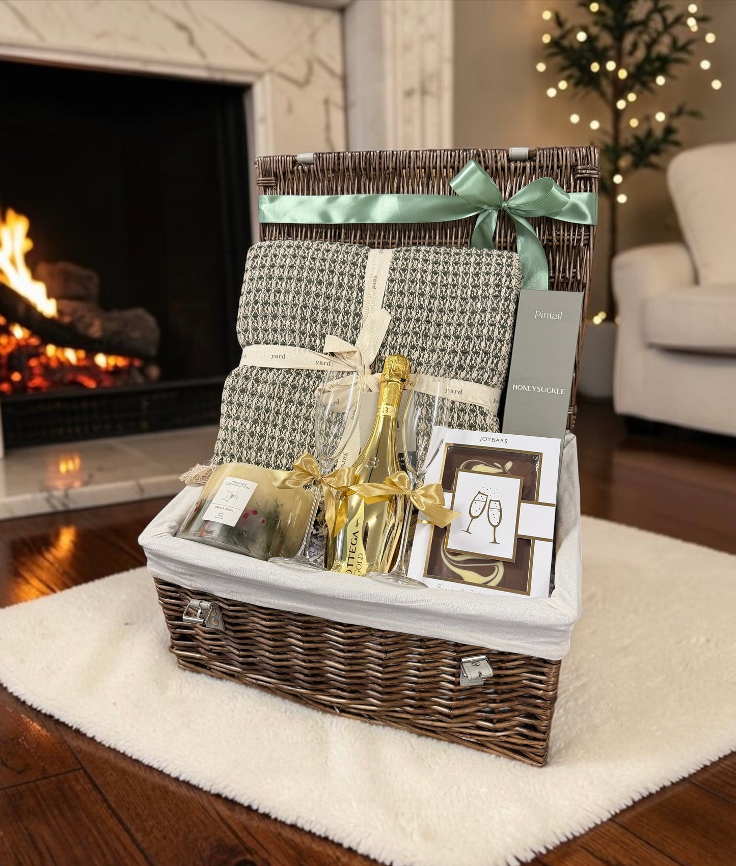 A wicker gift basket filled with a patterned throw blanket, champagne bottle, gold flute, and gift cards, set on a white rug in front of a lit fireplace in a cozy living room.