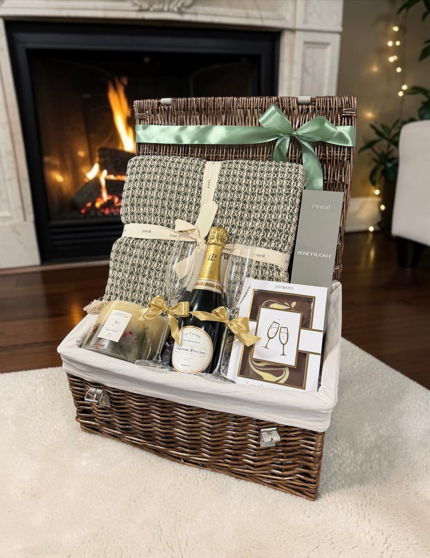 A wicker gift basket with a white fabric liner, filled with a patterned throw blanket, a bottle of champagne, a candle, and a champagne toast card, set in front of a lit fireplace.