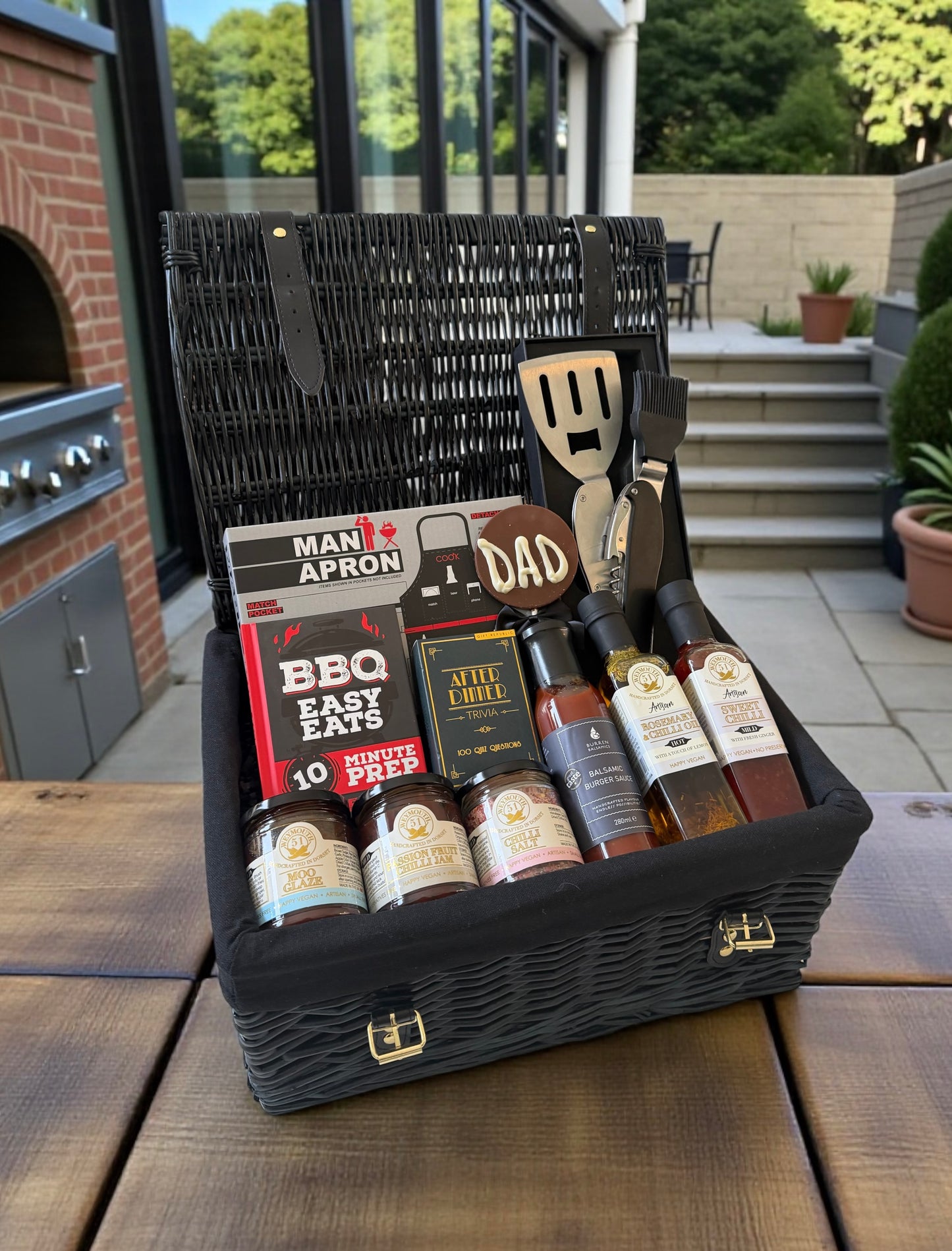 Dad’s BBQ hamper with BBQ apron, sauces, seasonings, personalised BBQ tool, recipe book, chocolate Dad lollipop, and optional drink cans.