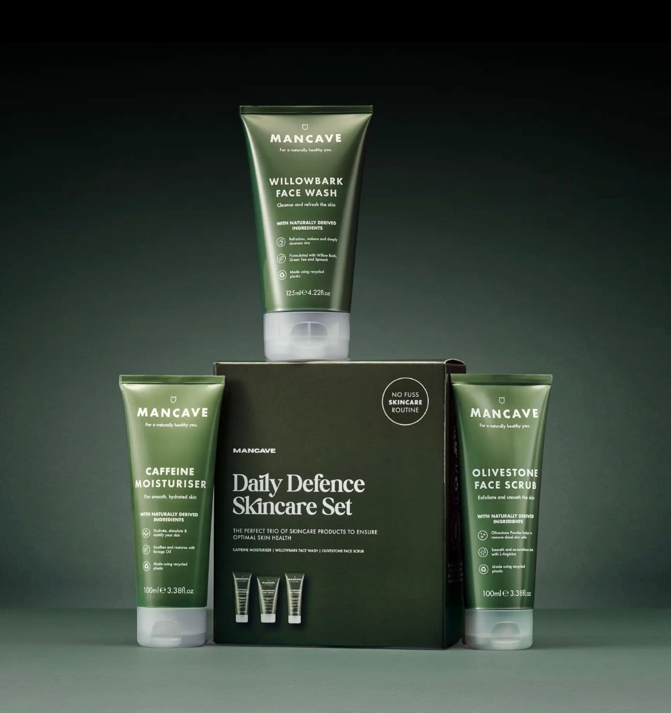 Mancave Daily Defence Skincare Set featuring a dark green box in the center with three matching green skincare tubes arranged around it: Caffeine Moisturizer, Willowbark Face Wash, and Olivestone Face Scrub, photographed on a dark gradient studio background.