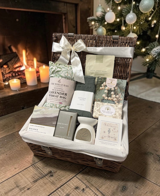 A wicker gift basket filled with assorted spa and home fragrance items, including a lavender steam bag, bath salts, soap, and a candle, arranged on a rustic wooden floor in front of a warm fireplace and a decorated Christmas tree.