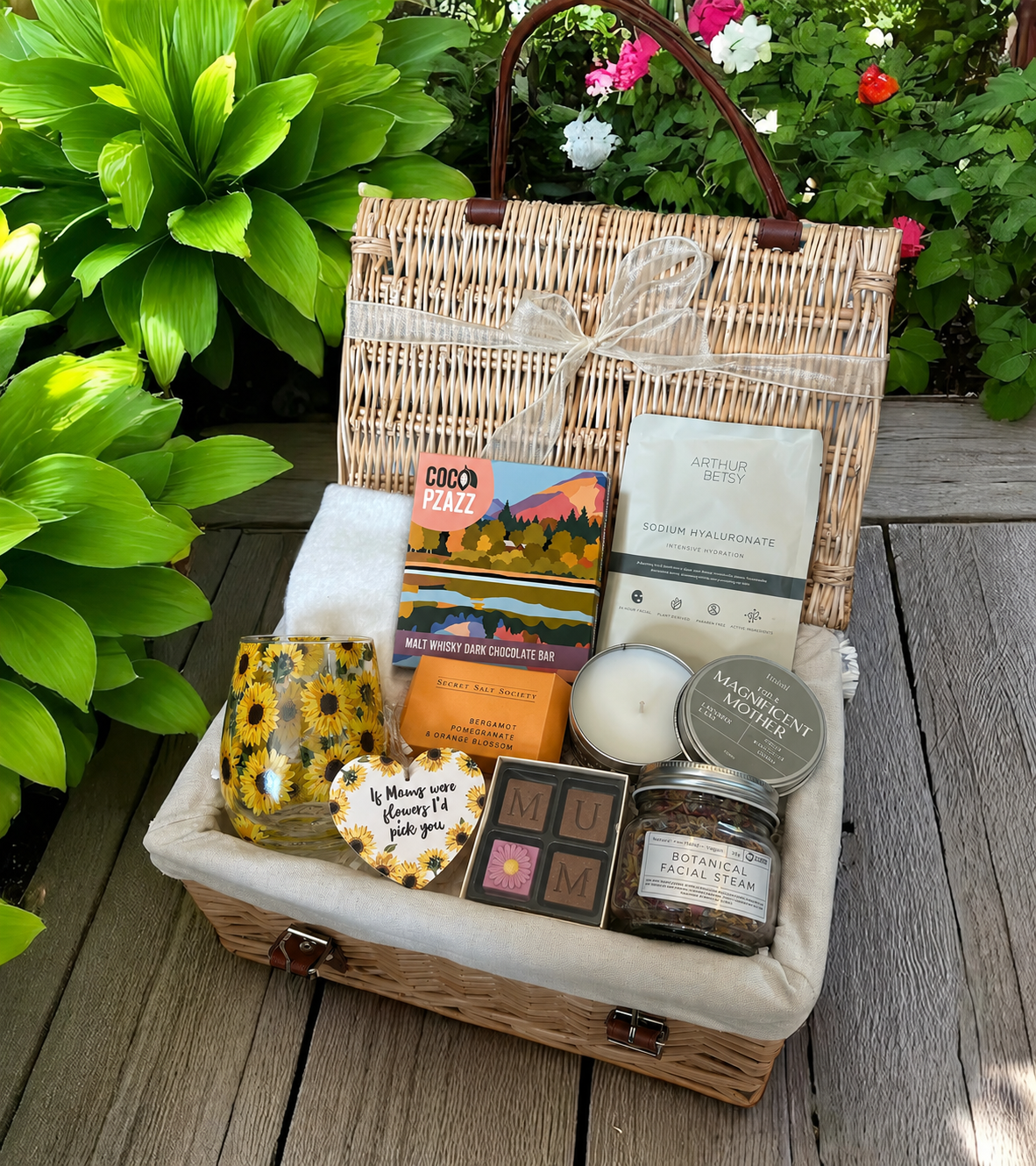 A wicker picnic basket filled with gift items, set on a wooden deck with green plants in the background.