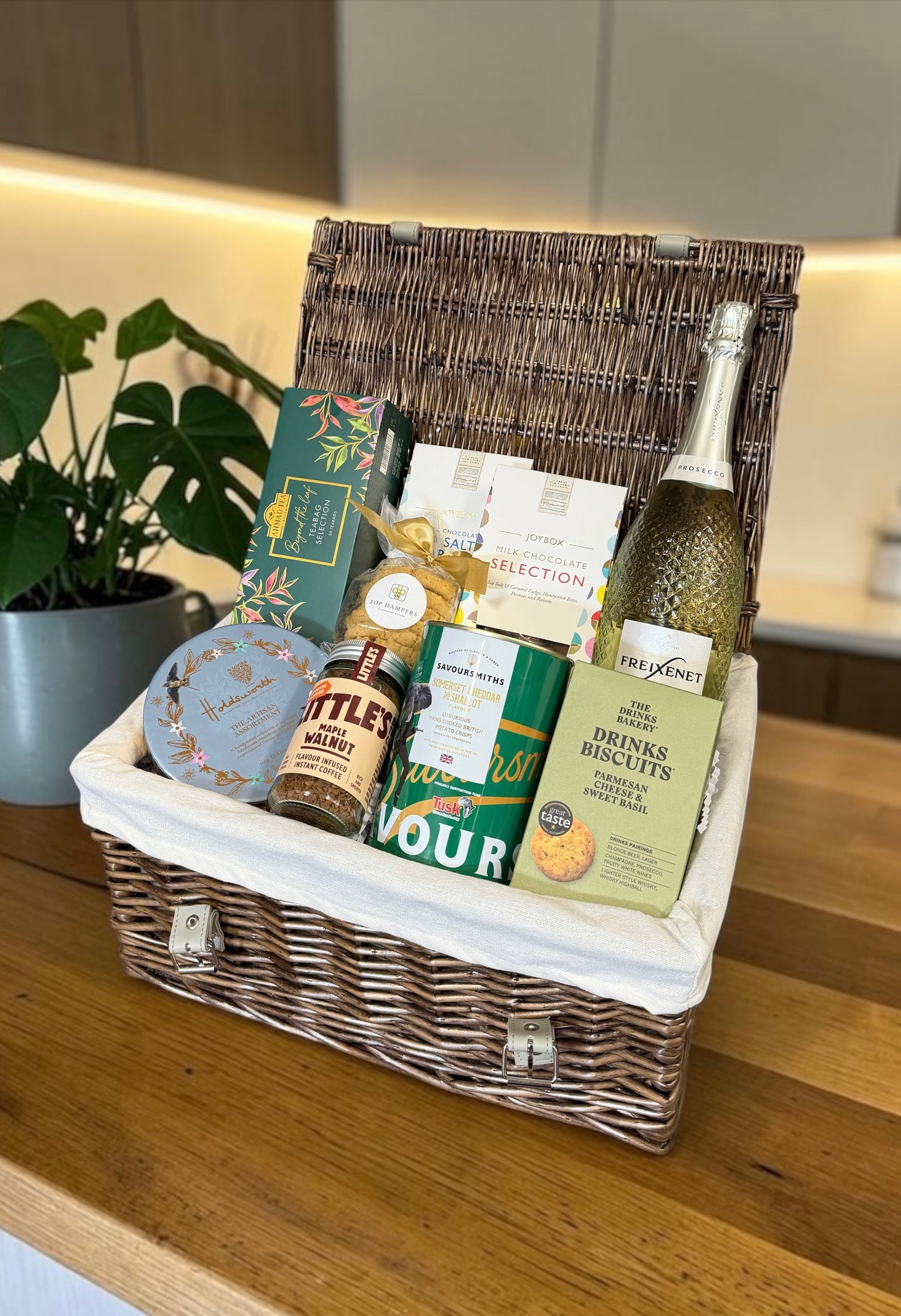 A wicker gift basket filled with assorted gourmet items, including a bottle of wine, crackers, biscuits, and packaged snacks, displayed on a wooden countertop in a kitchen setting.