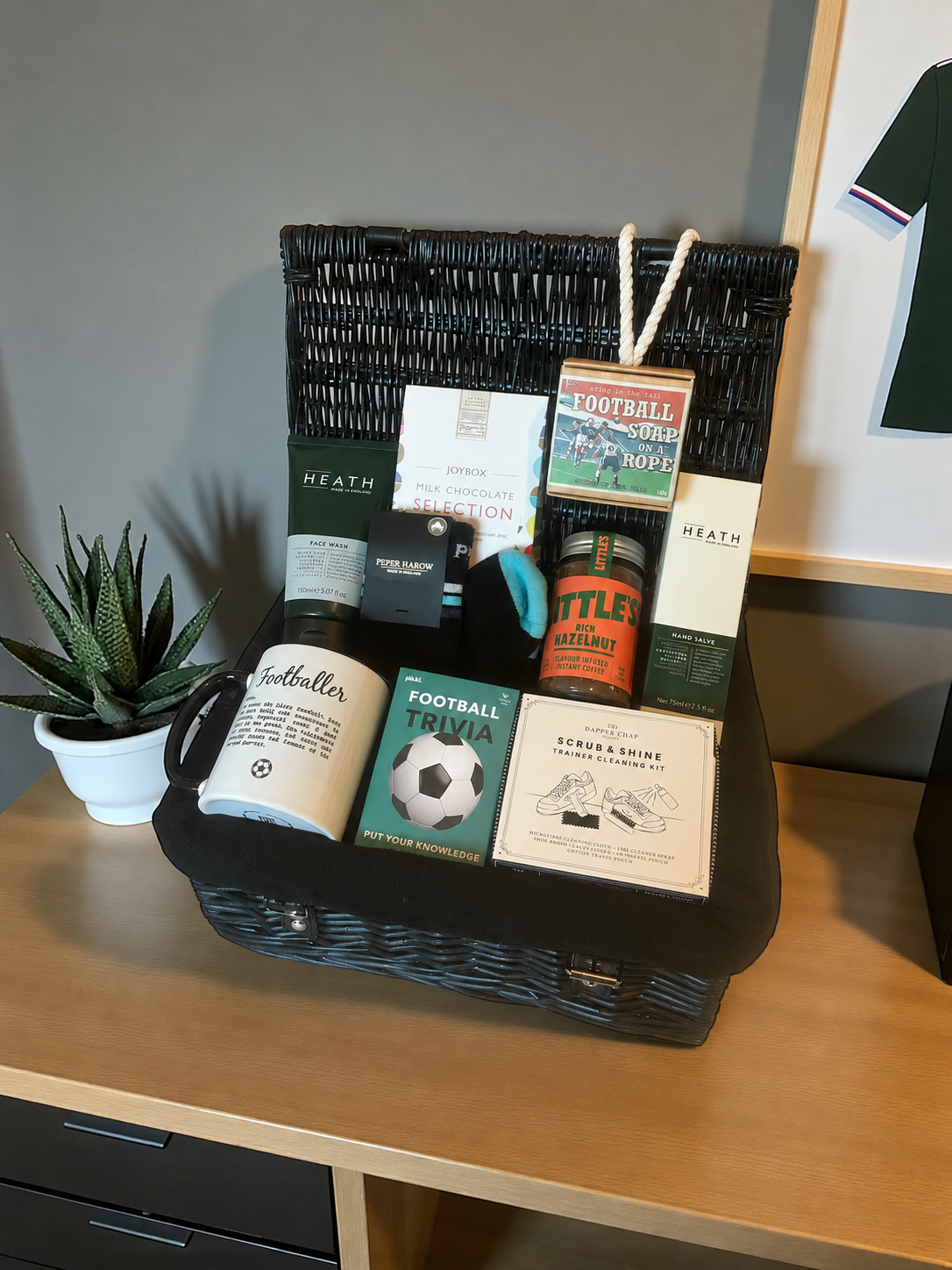 A black wicker gift hamper filled with assorted products including a mug, books, a candle, and packaged items, displayed on a wooden desk next to a small potted succulent.