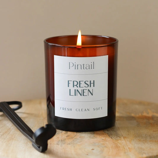 Fresh Linen Glass Candle with soothing floral and musk fragrance in amber jar on wooden surface.