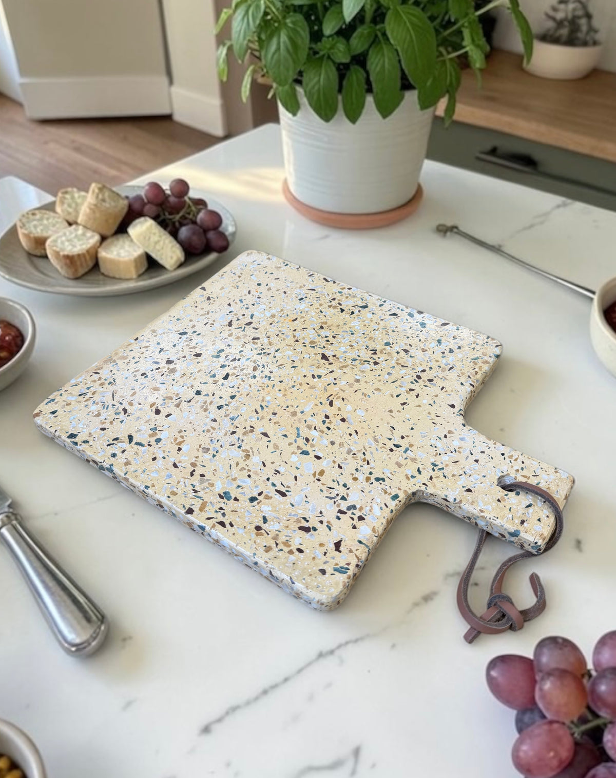 A rectangular terrazzo cheese board with a handle and hanging hook, styled on a white marble kitchen counter with food props and a potted plant in the background.