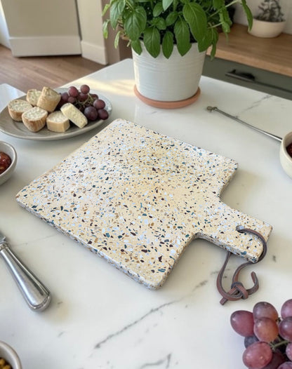 A rectangular terrazzo cheese board with a handle and hanging hook, styled on a white marble kitchen counter with food props and a potted plant in the background.
