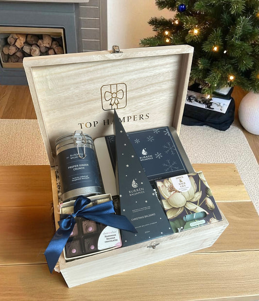 Gift hamper with chocolates, mulled wine gift set, candles, and other items in a wooden box, on a wooden surface with a Christmas tree in the background.