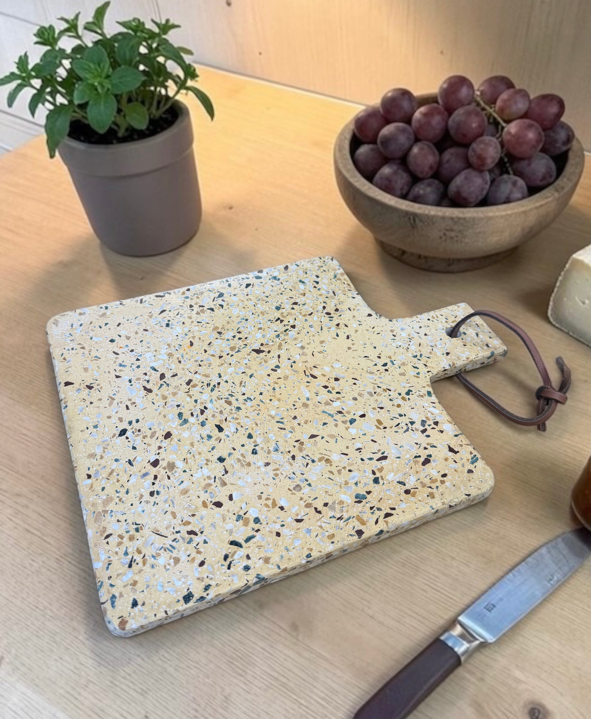 A rectangular terrazzo-style cutting board with a handle and dark brown cord tie, shown on a light wood table with a potted plant, grapes, cheese, and a knife nearby.