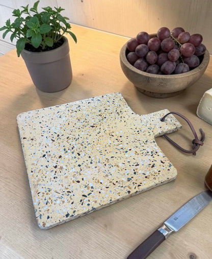 A rectangular terrazzo-style cutting board with a handle and dark brown cord tie, shown on a light wood table with a potted plant, grapes, cheese, and a knife nearby.