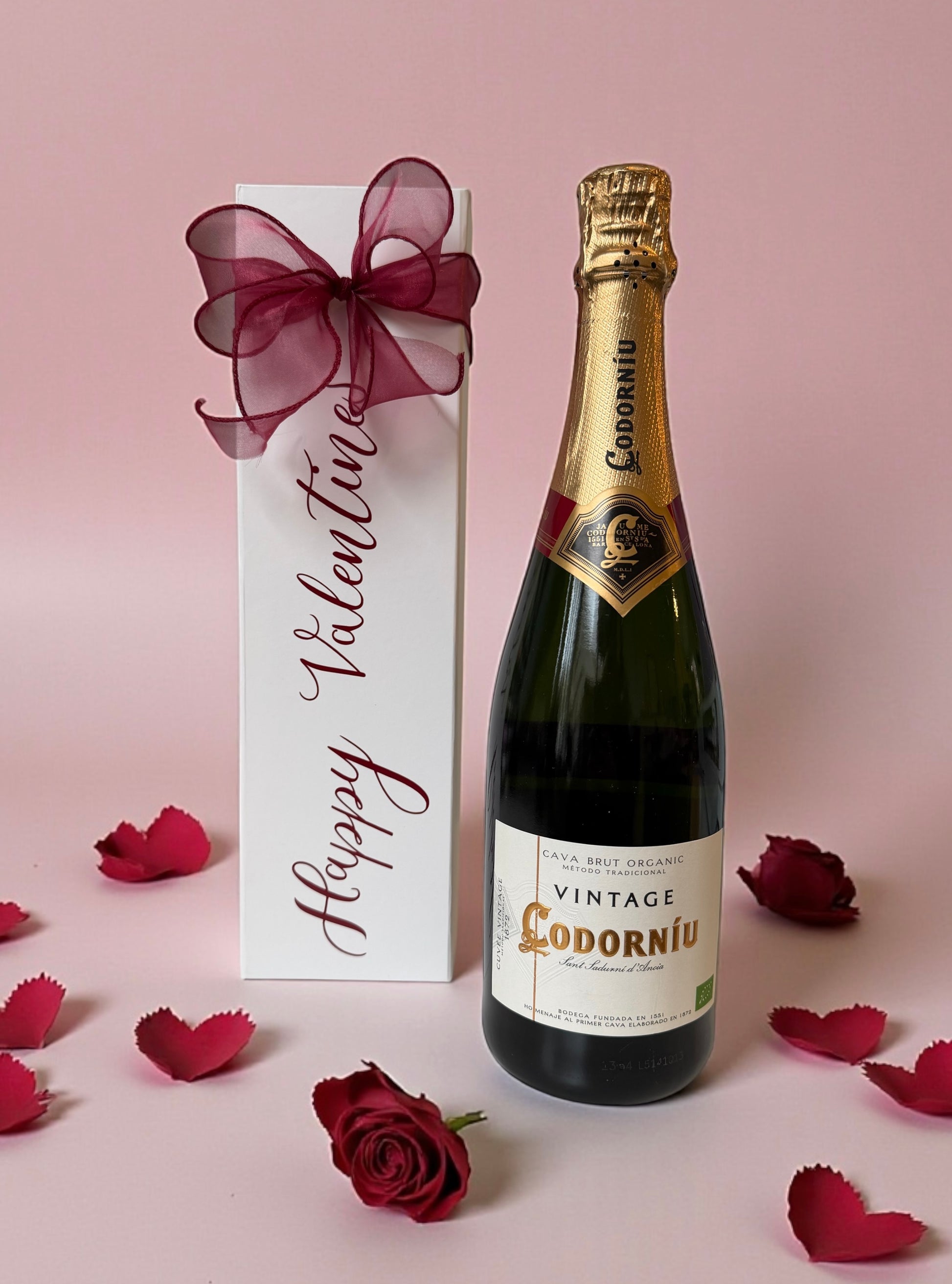 Bottle of Codorniu vintage champagne with a 'Happy Valentine's' gift box on a pink background.