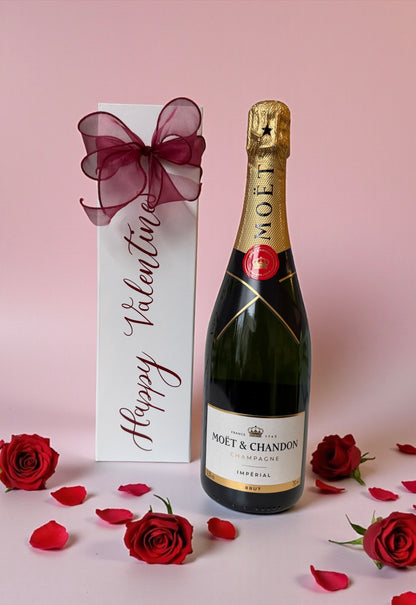 Moet & Chandon champagne bottle with a 'Happy Valentine's' gift box on a pink background with red roses.