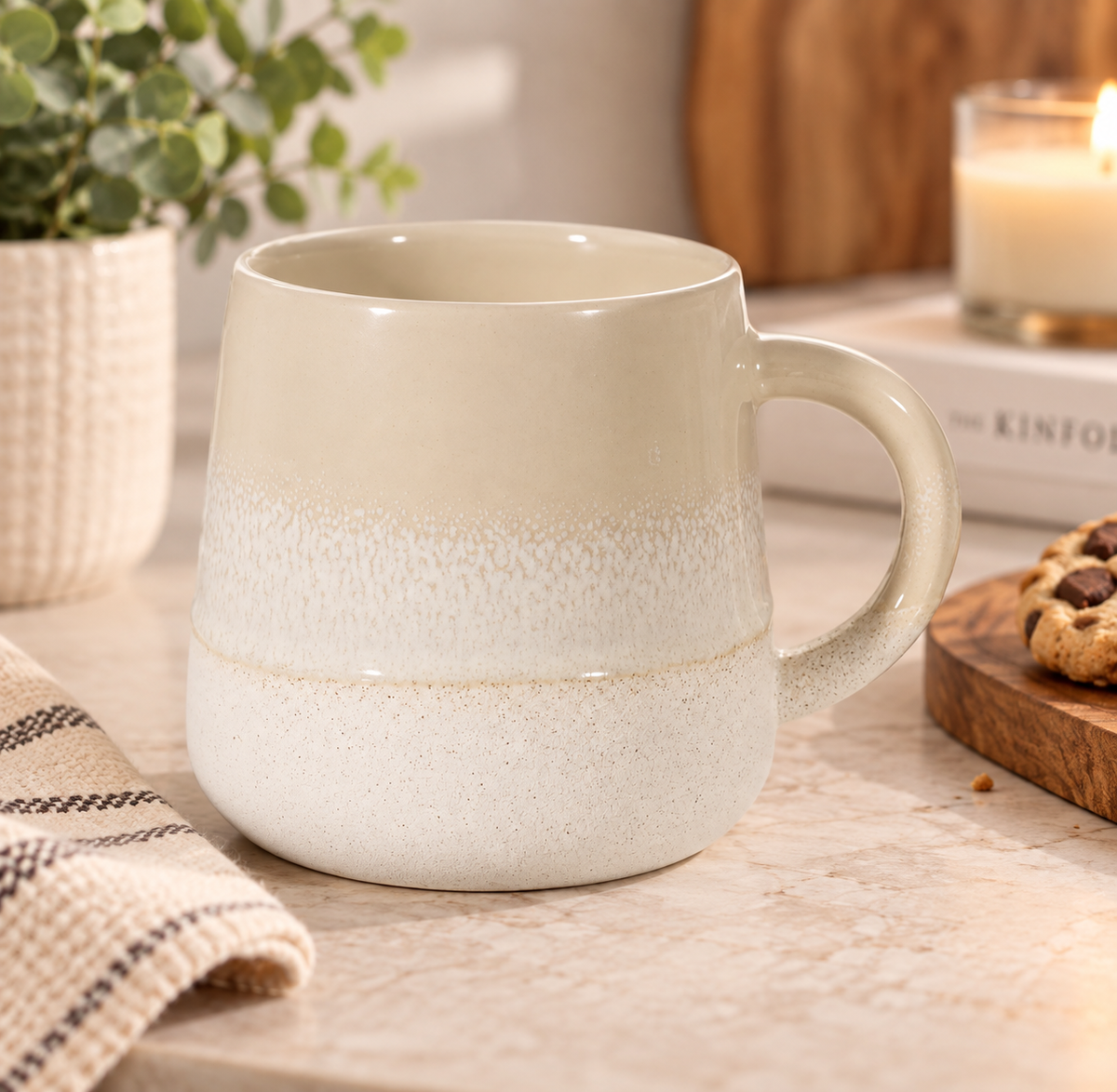 A ceramic mug with a smooth light beige upper half and a textured off-white speckled lower half, sitting on a wooden table with a plant, candle, and cookie in the background.