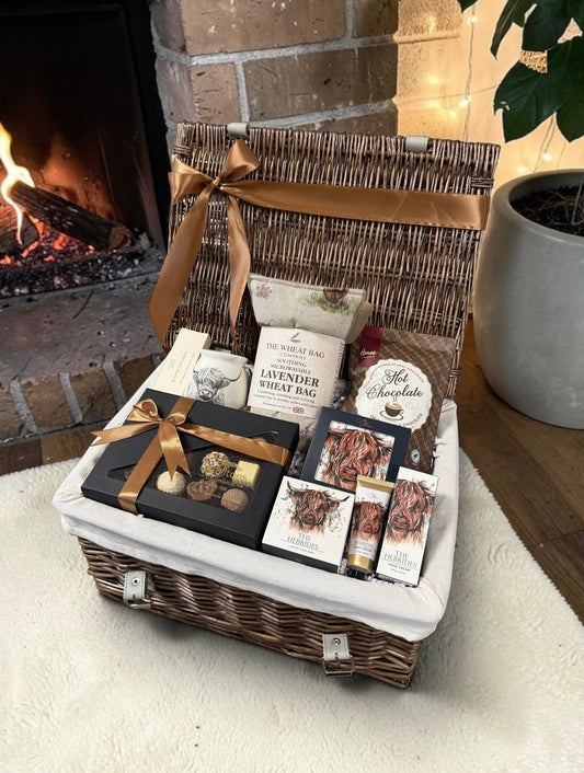 A wicker gift basket filled with assorted gourmet treats, chocolates, a lavender wheat bag, and a chocolate bar, styled in front of a warm fireplace.