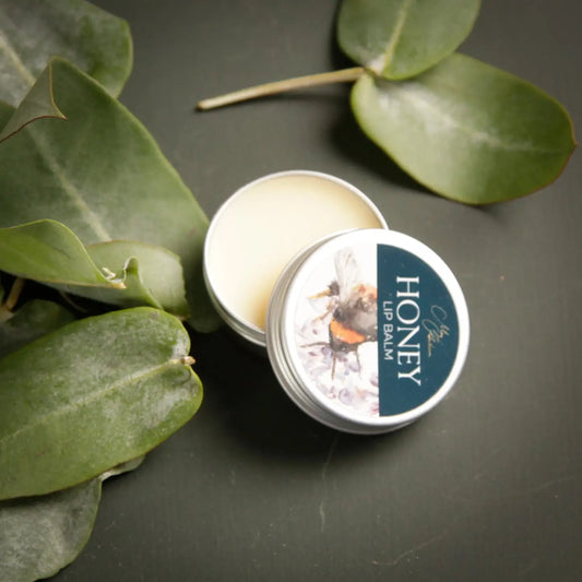 Honey Lip Balm with Bee Design in tin, surrounded by green leaves.