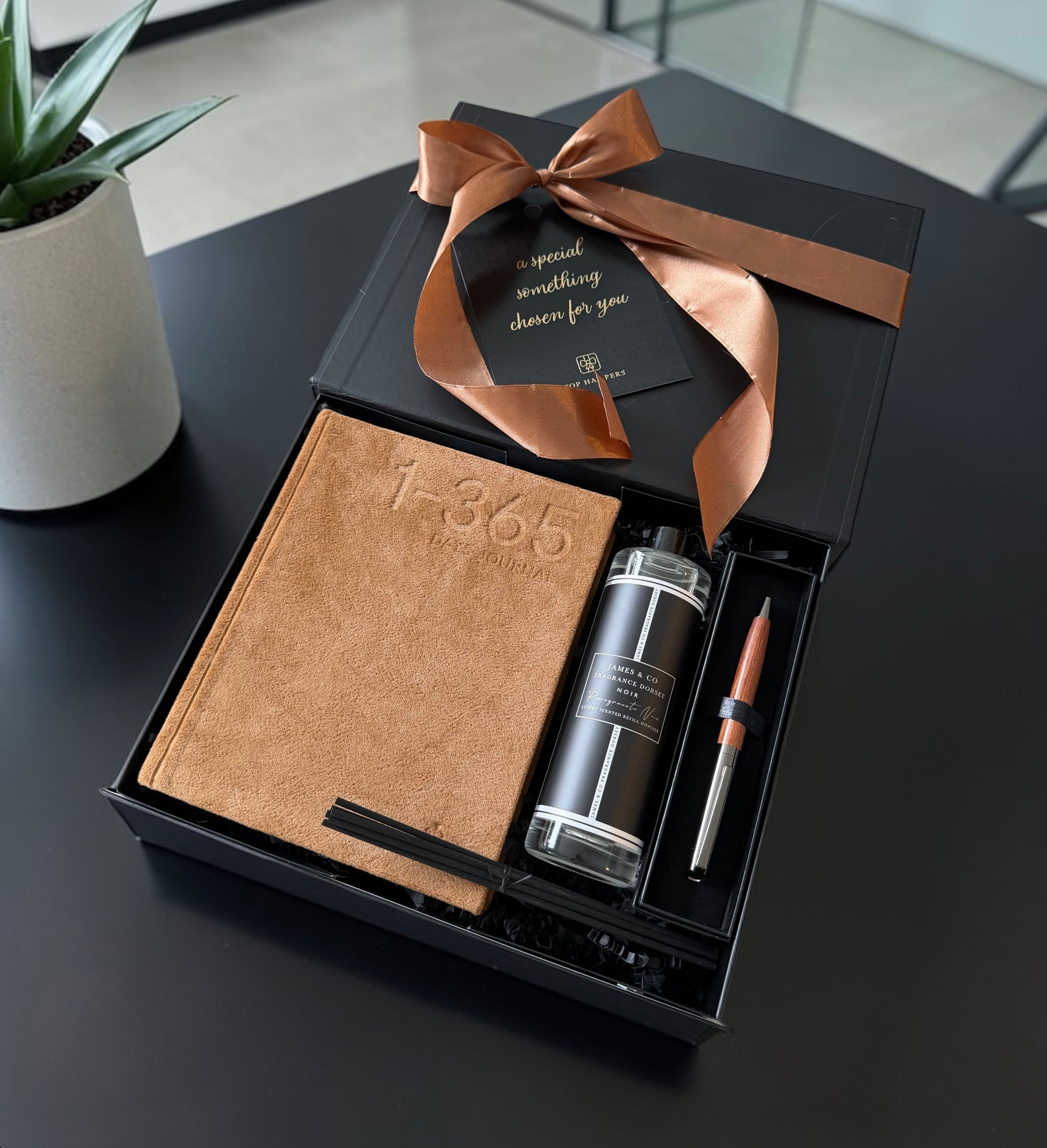 Gift set with a notebook, pen, and small bottle in a black box with a brown ribbon on a dark surface.