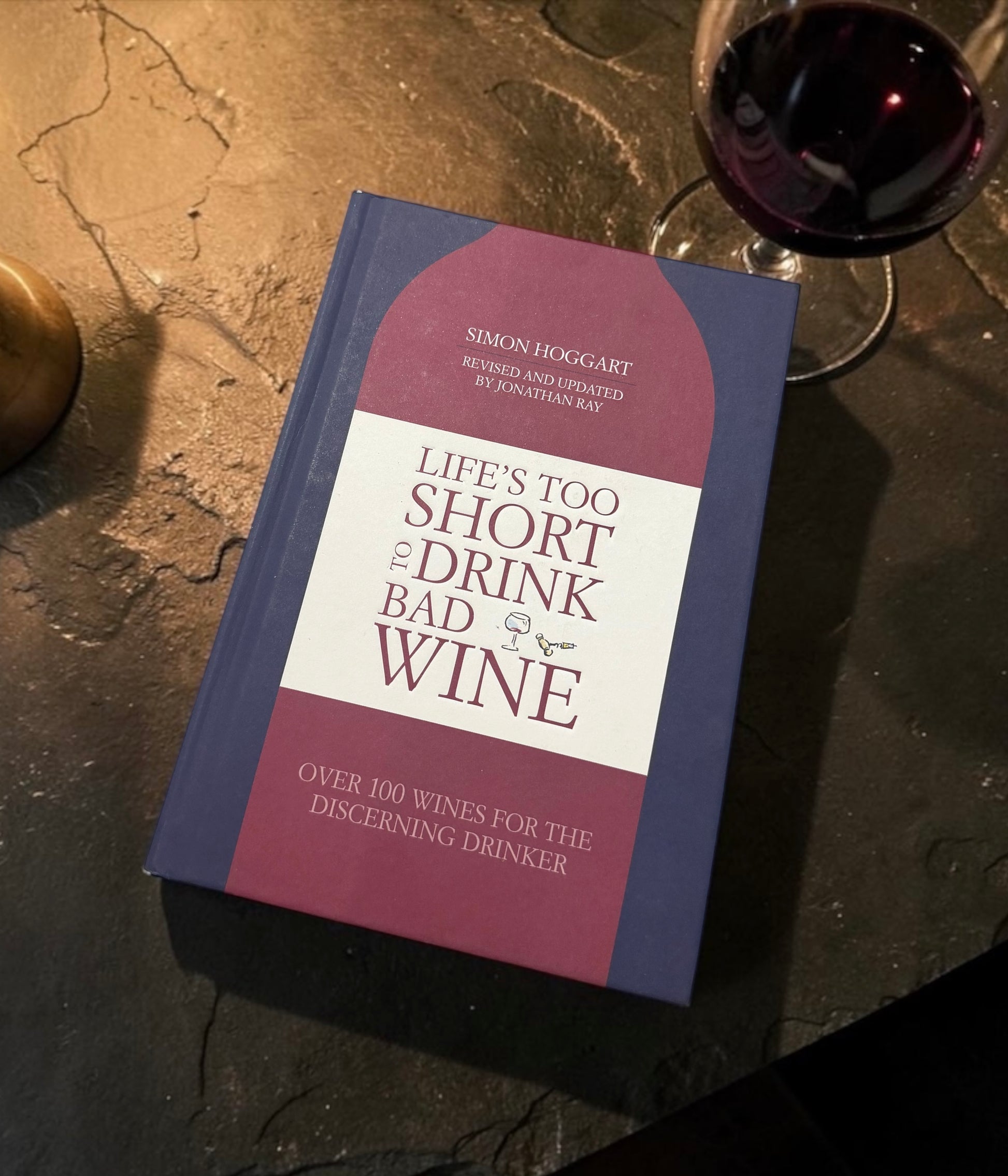 A hardcover book titled "Life's Too Short to Drink Bad Wine" by Simon Hoggart, placed on a dark stone surface next to a glass of red wine.