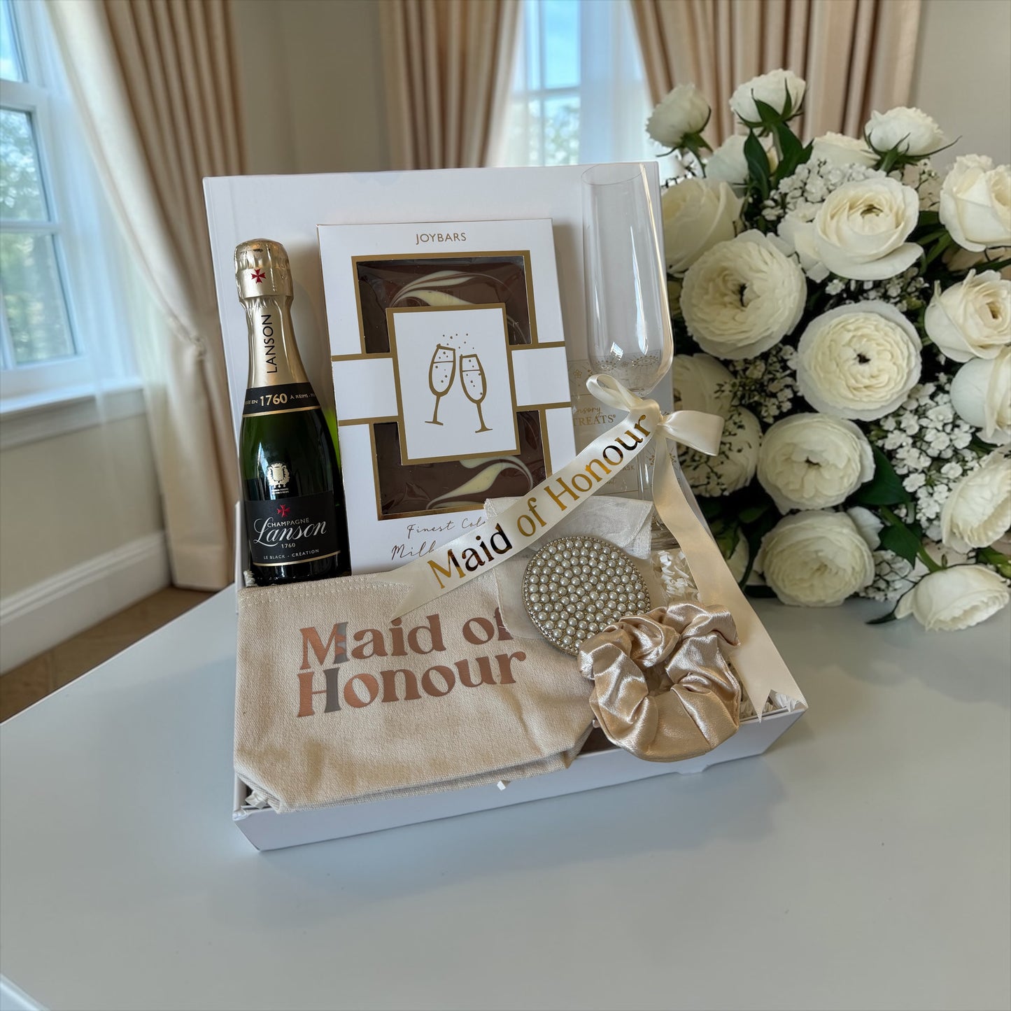 Gift basket with champagne, flowers, and 'Maid of Honour' items on a table.