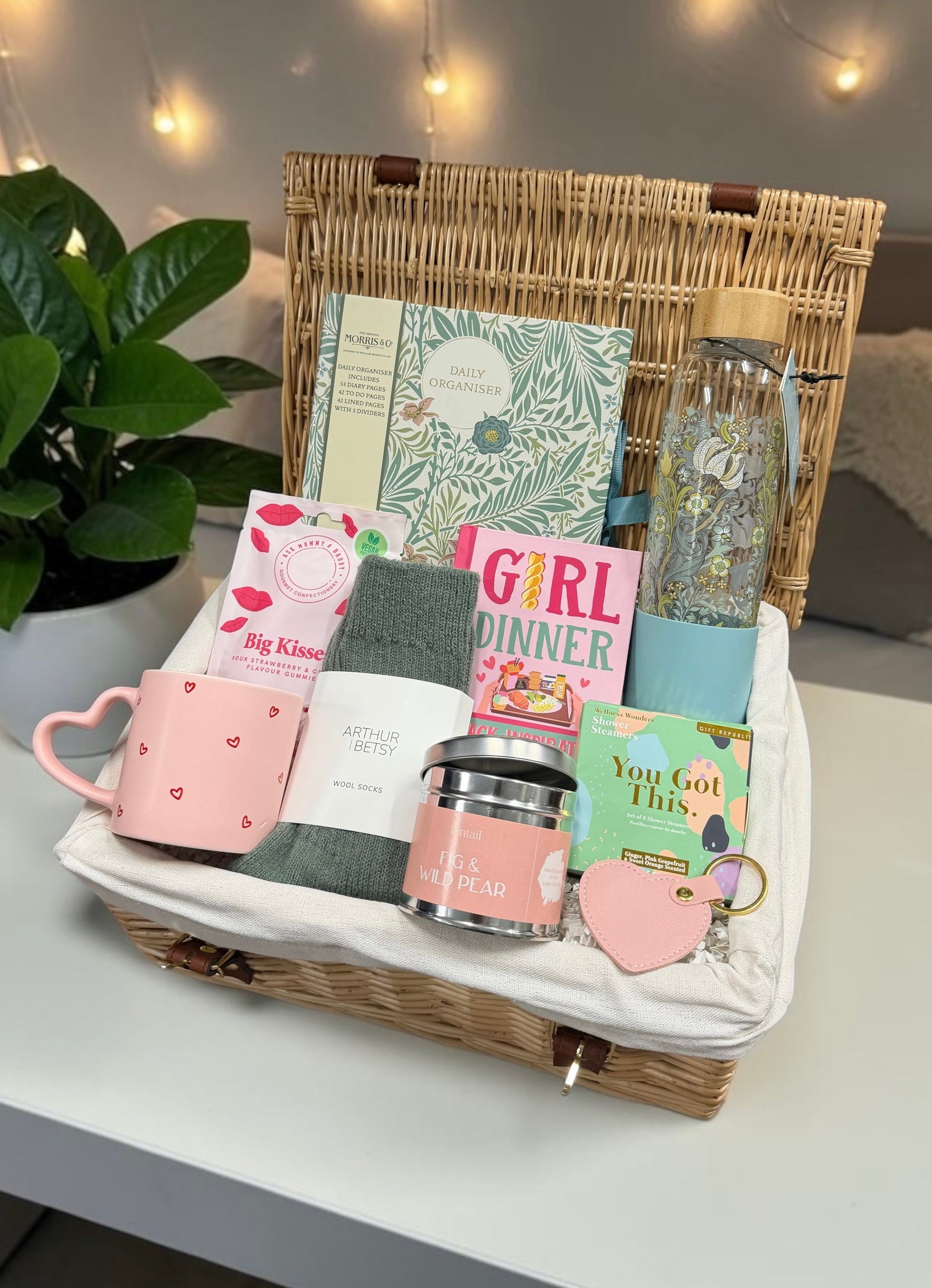 A wicker picnic-style gift basket arranged on a white table with a plant and warm string lights in the background. The basket contains a pink heart mug, a pink gift card, a green towel, a pink candle tin, a small book or notebook, a blue tumbler, a glittery water bottle, and other gift items.