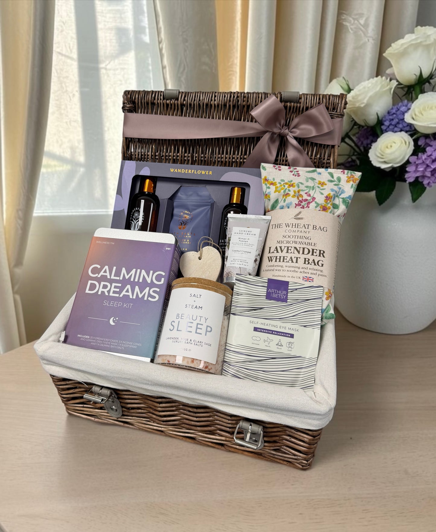 A wicker gift basket filled with wellness and relaxation items, including a purple 'Calming Dreams' sleep kit, a 'Be Well Sleep' box, lavender wheat bag, and bath products, set on a table next to a vase of white and purple flowers.