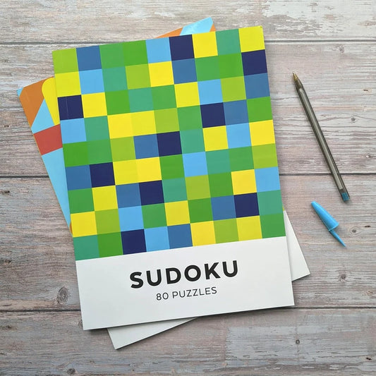 Sudoku - Adult Puzzle Book on a wooden table with colorful grid cover and pen.