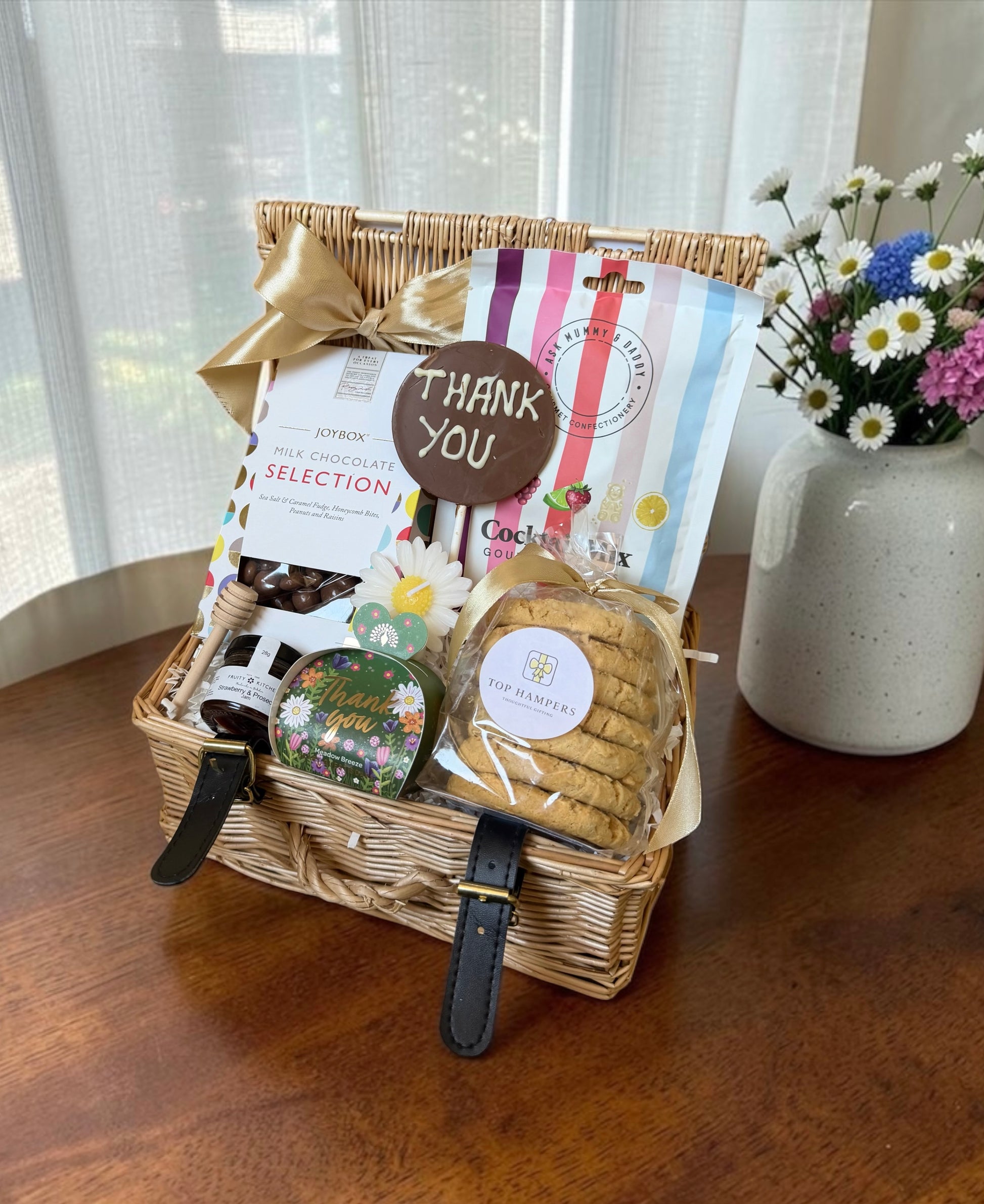 A thank you wicker gift basket filled with assorted gourmet treats, including a bag of chocolate selection, cookies, and a small thank you soap, tied with a gold ribbon and bow. The basket sits on a wooden table next to a vase of flowers.