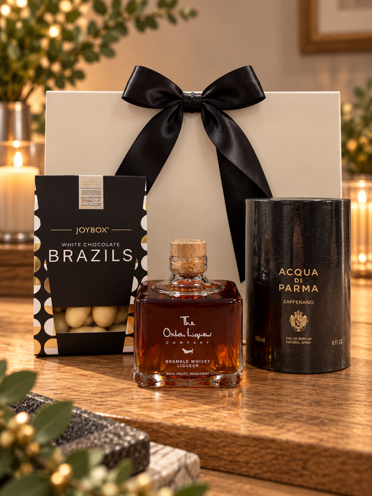 The Acqua di Parma Zafferano Gift Bag on a wooden table, with a box of Joybox white chocolate Brazils, a bottle of The Oxton Liqueur Company Bramble Whisky Liqueur and a 180ml Acqua di Parma Zafferano eau de parfum in its black tube, in front of a white gift bag tied with a black satin ribbon.