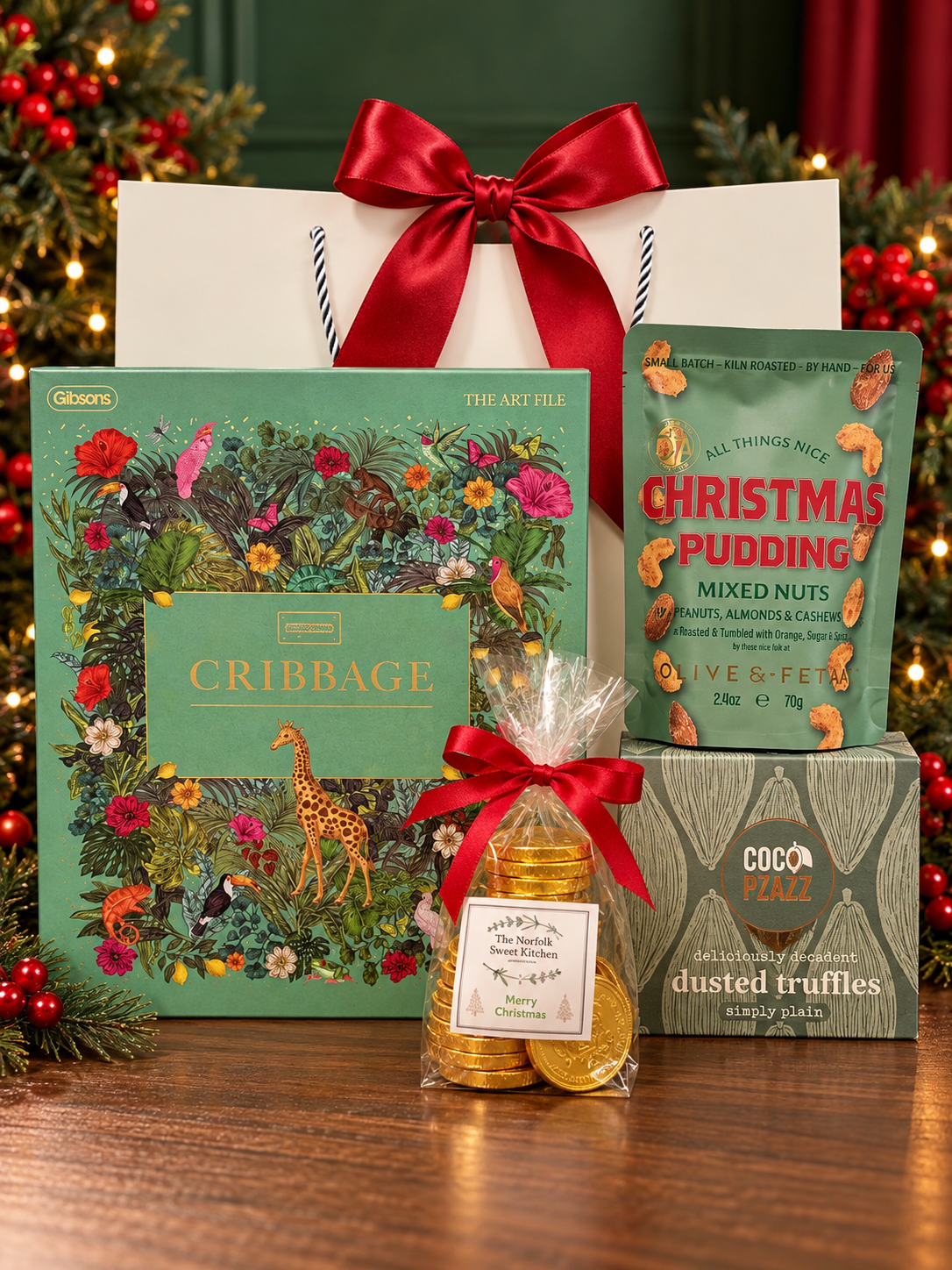 The Christmas Cribbage Gift Bag on a wooden table, with a Gibsons cribbage set in an illustrated jungle box, Olives Et Al Christmas Pudding mixed nuts, Coco Pzazz dusted truffles and a bag of gold chocolate coins, in front of a white gift bag tied with a satin ribbon.