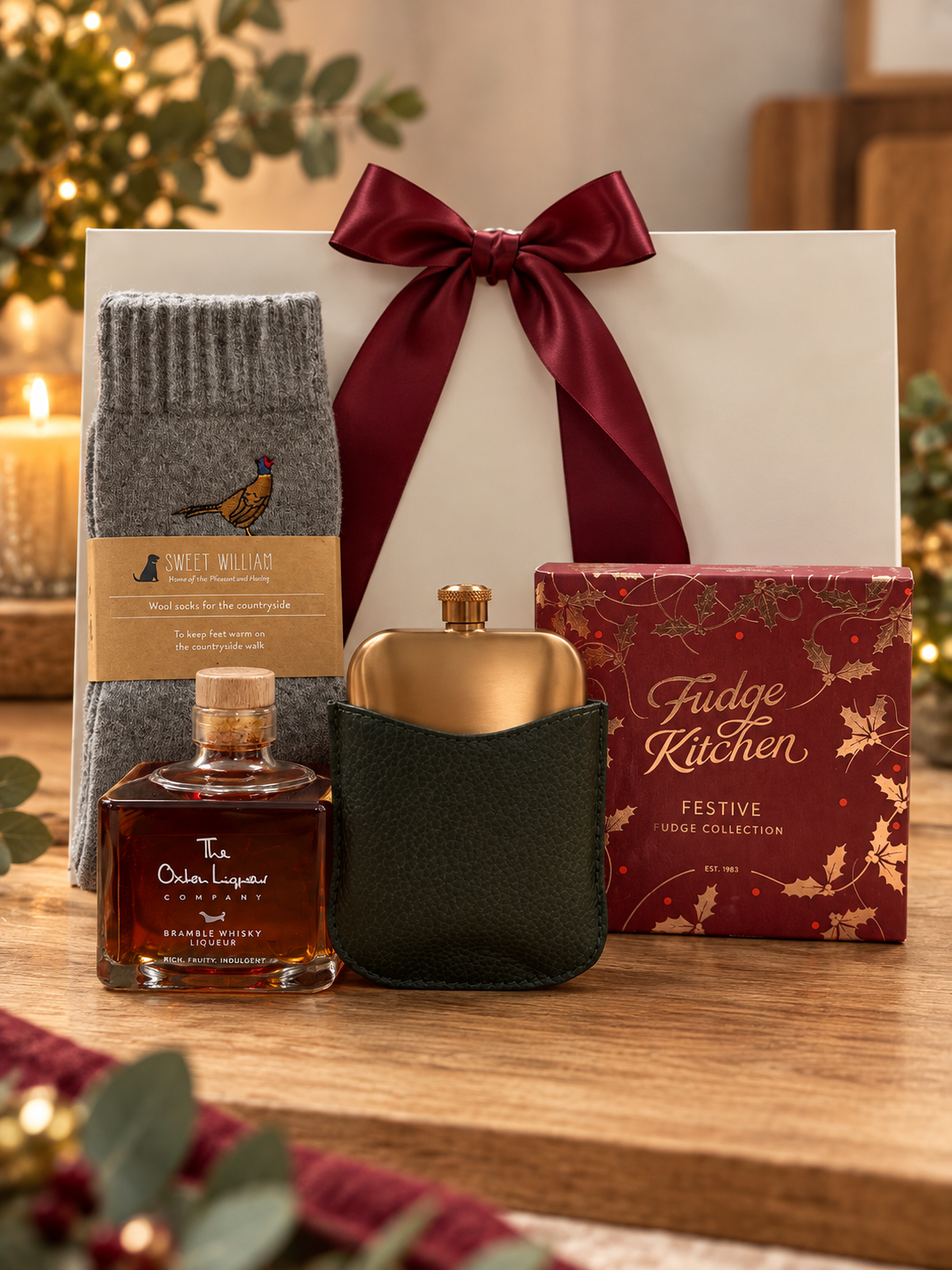 The Countryside Walk Gift Bag on a wooden table, with a pair of grey Sweet William wool socks with an embroidered pheasant, a bottle of The Oxton Liqueur Company Bramble Whisky Liqueur, a gold toned hip flask in a green pouch and a Fudge Kitchen Festive Fudge Collection, in front of a white gift bag tied with a burgundy satin ribbon.